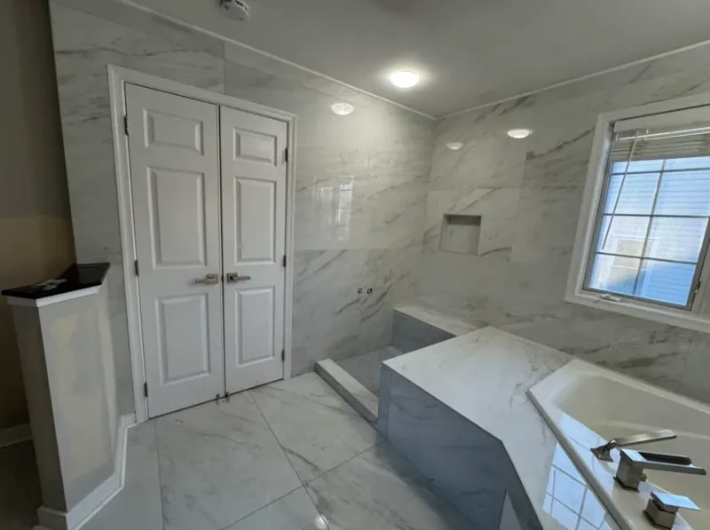 Wide bathroom view showing a tiled tub deck, white double doors, and marble-look wall and floor surfaces